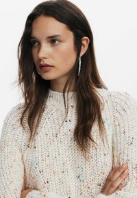 Maglione lavorato a maglia in crema con macchie colorate, caratterizzato da un collo alto e maniche lunghe. Il modello indossa orecchini a cerchio texturizzati.