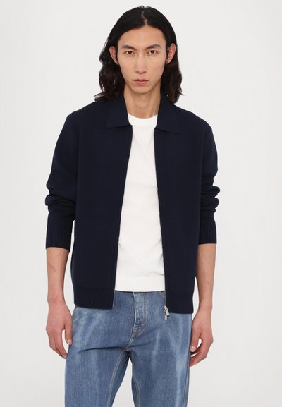 Cardigã azul marinho com fecho éclair, com gola e punhos canelados, feito de tecido tricotado. Usado com uma t-shirt branca e jeans azul de ganga.