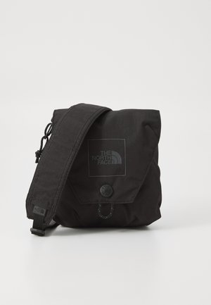 Sac à épaule en nylon noir avec rabat, présentant un patch logo, une sangle réglable et un système de fermeture à pression. Surface texturée avec des détails cousus.