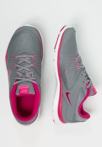 Zapatillas deportivas grises con acentos en rosa, parte superior de malla y suela blanca. Incluyen forro interior rosa, cordones y el logo de Nike en el lado.