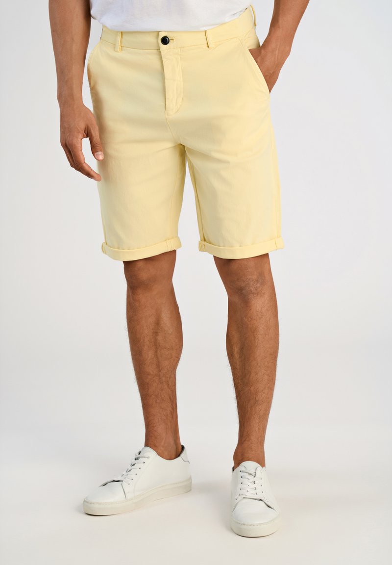 Shorts jaunes clair en coton, avec des ourlets retroussés, des poches avant et une fermeture par bouton, associés à des baskets blanches.