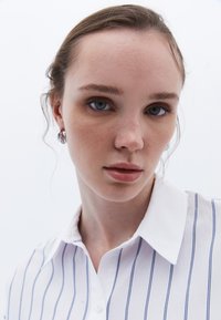 Camisa blanca de botones con rayas verticales azules, con un cuello puntiagudo. El modelo tiene piel clara y lleva aros pequeños.