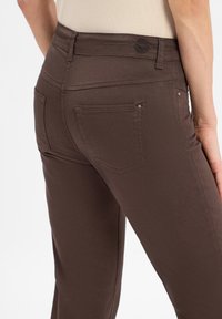 Nahaufnahme einer Person, die enge, dunkelbraune Jeans mit Gesäßtaschen und Gürtelschlaufen trägt, die Taille und Oberschenkel zeigt.