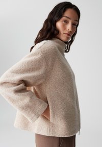 Giacca in misto lana beige con colletto alto, maniche a tre quarti e finitura texturizzata. Presenta una zip laterale e una vestibilità rilassata.