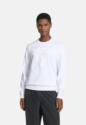 Junger Mann trägt ein weißes Sweatshirt mit erhöhtem GUESS-Logo und dunkelgrauen Hose, steht vor einem einfarbigen hellen Hintergrund.