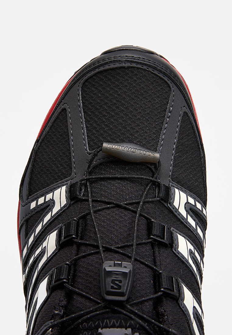 Schwarzer Sportschuh mit strukturiertem Obermaterial, weißen Akzenten und roter Außensohle. Verfügt über elastische Schnürsenkel und ein Metall-Branding-Tag.