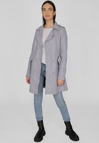 Lichtgrijze trenchcoat met een dubbelgekeerd ontwerp, grote knopen en schuine zakken, gecombineerd met lichtblauwe jeans en zwarte enkellaarsjes.