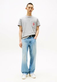 Camiseta gris de cuello redondo con logo rojo en el bolsillo, combinada con jeans anchos azules y zapatillas blancas, de pie contra un fondo claro.
