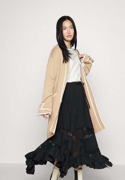 Beige gebreide cardigan met witte rand, gelaagd over een witte top en een zwarte, geplooide rok met ruches en doorschijnende panelen.