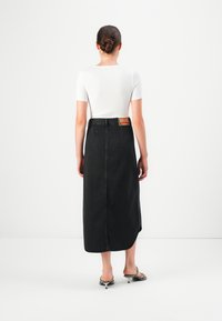 Kvinne med håret i en knute iført hvit kortermet topp, svart midi denimskjørt med etikett, og svarte hælsandaler, vendt bort fra kameraet mot hvit bakgrunn.