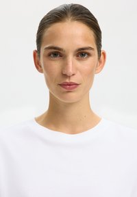 Femme de face avec les cheveux bruns coiffés en arrière, portant un t-shirt blanc uni à col rond sur un fond blanc.