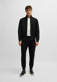 Zwarte gezipte jas met hoge kraag, gecombineerd met zwarte trekkoordbroek en zwarte sneakers. Glad materiaal met een comfortabele pasvorm.