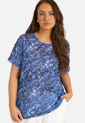 Ung kvinde med langt brunt hår iført en blå mønstret kortærmet skjorte og hvide bukser, stående med den ene hånd i lommen.