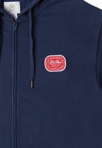 Námornícky modrá mikina na zips vyrobená z mäkkého materiálu, s červeným a bielym logom na hrudi a s nastaviteľnými šnúrkami na kapucni.