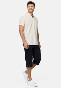 Camisa de botón de manga corta en color crema, pantalones cortos de carga en color marino y zapatillas blancas. La camisa presenta un cuello clásico y un corte relajado; los pantalones cortos tienen bolsillos.