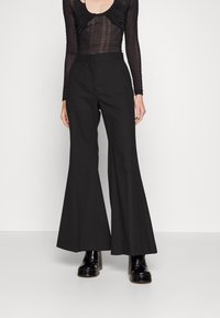 Person som bär svarta utsvängda byxor, svart genomskinlig långärmad topp och svarta plattformsskor, stående mot en vit bakgrund.