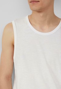 Witte mouwloze katoen T-shirt met een ronde hals en subtiele textuur, met een klein, licht logo in de linkerbenedenhoek.