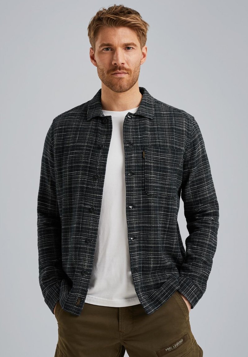 Chemise à boutons en design carreaux noir et gris, avec un col, des manches longues et un tissu texturé. Portée sur un t-shirt blanc.