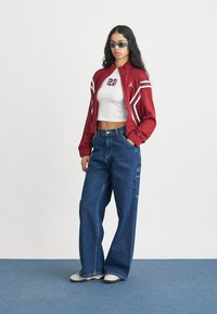 Rdeča bomber jakna z belimi črtami, kratka bela majica brez rokavov z barvito številko 23 in ohlapne modre jeans hlače. Stilska sončna očala dopolnjujejo videz.