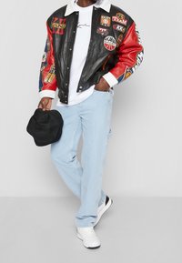 Giacca bomber in pelle nera con maniche rosse, decorata con varie toppe ricamate. T-shirt bianca, jeans azzurri e cappello a secchiello nero.