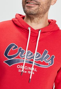 Czerwona bawełniana bluza z kapturem z białymi sznurkami, dużym logo "Creeks" w granatowym kolorze oraz napisem "Original Since 1994" pod logo.