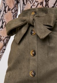 Jupe en daim vert olive avec un détail de nœud à l'avant et quatre boutons à motif marbré. Le tissu possède une finition douce et texturée.