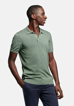 Jonge man die glimlacht en naar rechts kijkt, gekleed in een lichtgroene korte mouwen polo en donkerblauwe broek, met één hand in zijn zak.