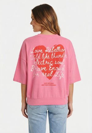 Donna che indossa una camicia rosa oversize con un grande cuore rosso e testo bianco scritto a mano sul retro, abbinata a jeans azzurri.