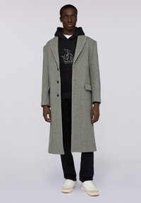 Šedý vlněný kabát s límečkem, jednobarevný design a dvěmi předními kapsami, nošený přes černou mikinu a tmavé kalhoty s bílými teniskami.