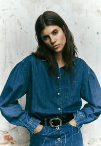 Camicia di denim con tasca, design con bottoni e maniche voluminose, abbinata a una cintura in pelle nera con fibbia rettangolare.