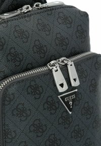 Black patterned backpack with textured synthetic material, silver zippers, and a triangular black logo patch featuring "GUESS."