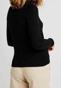 Schwarzer Strickpullover mit taillierter Passform, langen Ärmeln und gerippten Bündchen. Kombiniert mit hellbeigen Hosen, von hinten gezeigt.