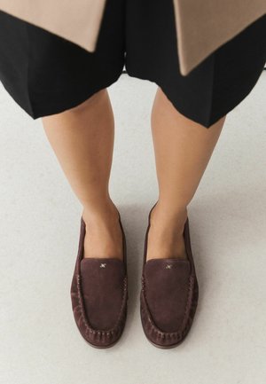 Femme portant des mocassins en daim brun foncé et une jupe noire, debout sur un sol clair.