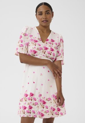 Mujer de cabello oscuro vestida con un vestido blanco con estampado floral rosa, mangas cortas abullonadas y escote en V, de pie frente a un fondo claro.