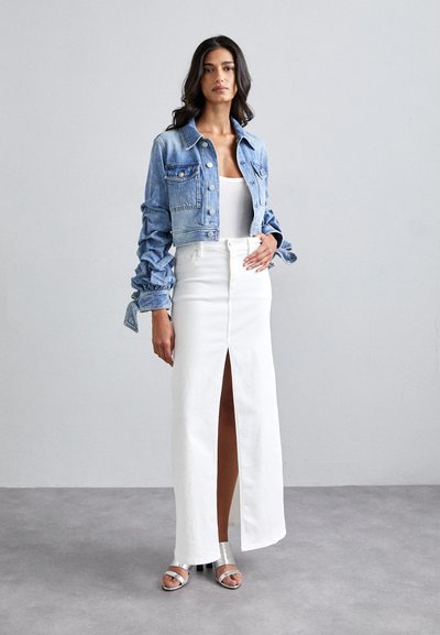Chaqueta de mezclilla corta con detalles de botones, combinada con una falda blanca de talle alto que presenta una abertura frontal. Sandalias de tacón plateadas completan el look.
