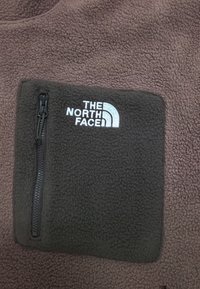 Dunkelbraunes Fleece-Kleidungsstück mit Reißverschlusstasche und gesticktem weißen "The North Face"-Logo auf der Tasche.