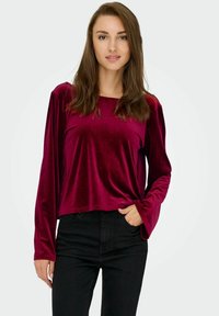 Burgundy sammet långärmad topp med en lös passform och vida ärmar, i kombination med svarta tajta jeans, som visar en mjuk, texturerad yta.