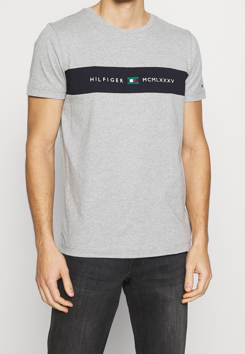 Camiseta de algodón gris con una franja horizontal negra que presenta el texto "HILFIGER" y "MCMLXXXV", junto a un logo de bloques de color. Mangas cortas.