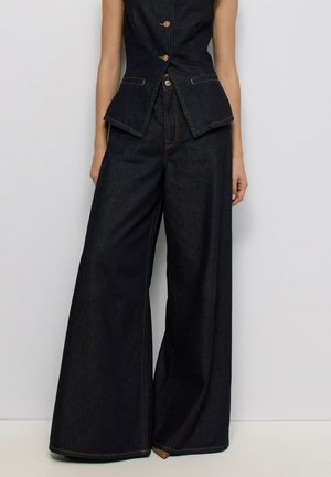 Mujer con pantalones vaqueros oscuros de pierna ancha y chaleco sin mangas a juego con botones, sobre fondo blanco.