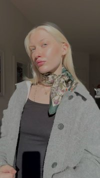 Grijze wollen jas met zwarte knopen over een zwart shirt, met een groene sjaal met bloemenprint gestrikt rond de hals, gladde textuur.