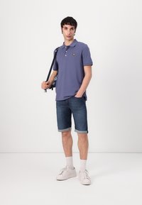 Šviesiai violetinė polo marškinėliai su geltonu logotipu, derinami su tamsiais džinsiniais šortais ir baltais sportbačiais. Modelis laiko raštuotą kuprinę.