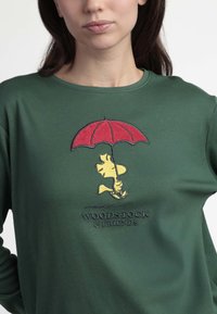 Camisa de manga larga verde con un pájaro amarillo bordado sosteniendo un paraguas rojo. Texto "WOODSTOCK & FRIENDS" en un color oscuro debajo del diseño.