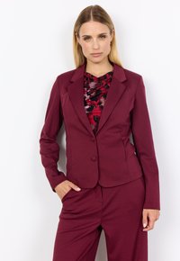 Frau mit glattem blonden Haar, die einen bordeauxfarbenen Anzug und ein floral gemustertes Oberteil trägt, steht vor einem weißen Hintergrund mit einer Hand in der Tasche.