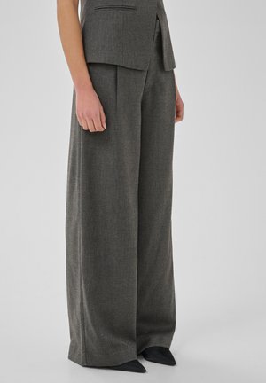 Personne portant un pantalon gris à jambes larges et un gilet assorti, avec des chaussures noires pointues visibles, debout devant un fond uni clair.