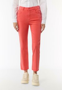 Coral pink denim pants with a slim fit, five-pocket design, and a subtle sheen. Paired with light-colored sneakers.
