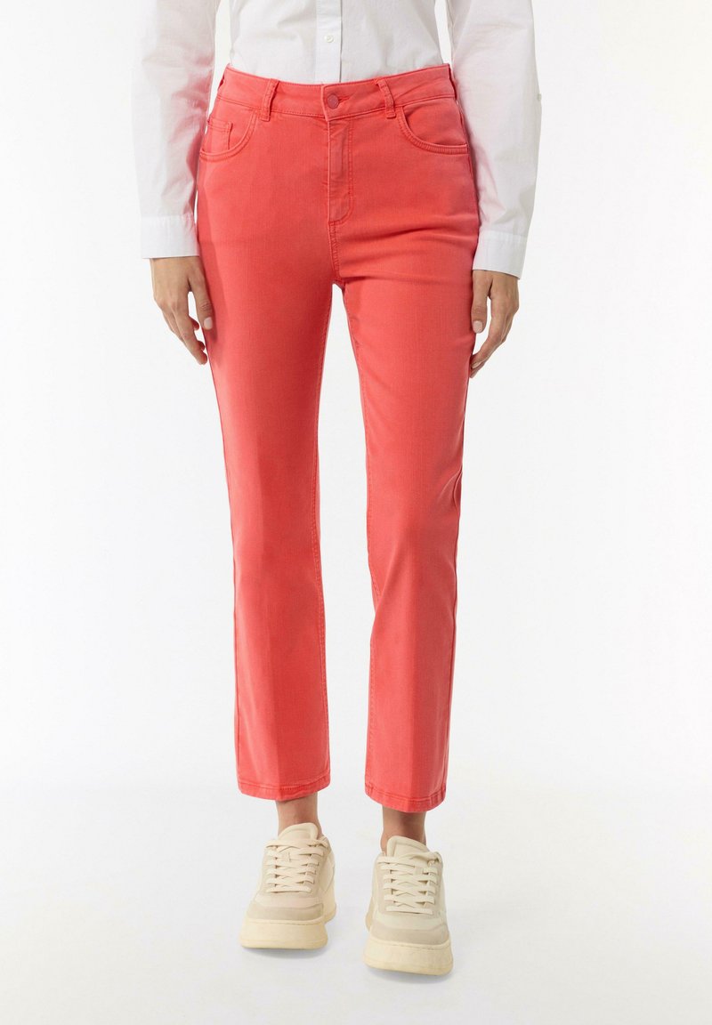 Coral pink denim pants with a slim fit, five-pocket design, and a subtle sheen. Paired with light-colored sneakers.