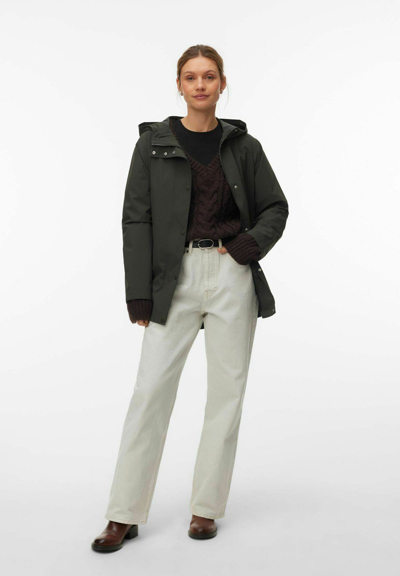 Femme portant une veste à capuche vert foncé, un pull en maille torsadée marron foncé, un pantalon écru, une ceinture noire et des bottes marron, debout avec une main dans la poche.