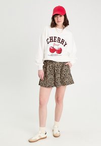 Sweatshirt blanc avec graphique de cerise et texte, associé à un short à imprimé léopard noir. Une casquette rouge et des baskets blanches complètent la tenue.