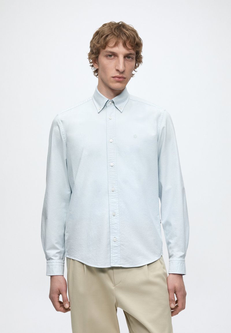 Jeune homme aux cheveux bouclés clairs portant une chemise bleu clair boutonnée et un pantalon beige, debout devant un fond blanc uni.