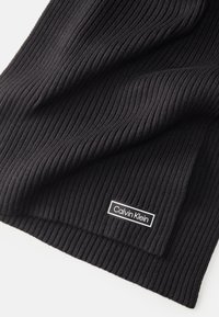 Écharpe tricotée noire à côtes avec un patch logo Calvin Klein noir et blanc cousu. Texture douce avec des rayures verticales régulières.